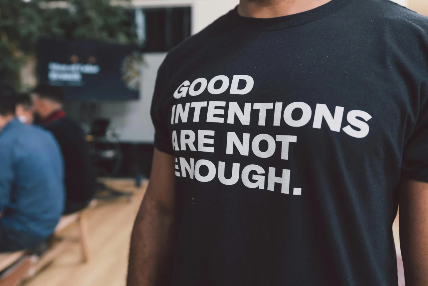 A man wearing a black t-shirt emblazoned with the words "GOOD INTENTIONS ARE NOT ENOUGH." in bold white print.