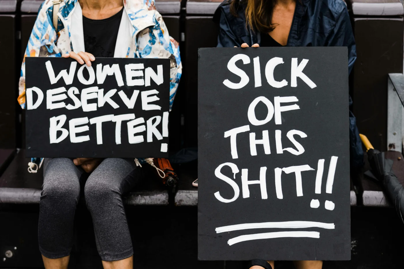 Two women sitting next to each other and holding signs at the Brisbane Women’s March 4 Justice, King George Square, Brisbane, Australia. One sign reads "WOMEN DESERVE BETTER!" and the other reads "SICK OF THIS SHIT!!".