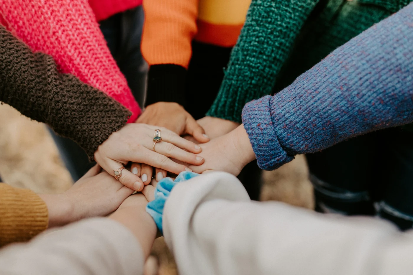 A close-up of the arms of a group of women in colourful jumpers stacking their hands together.