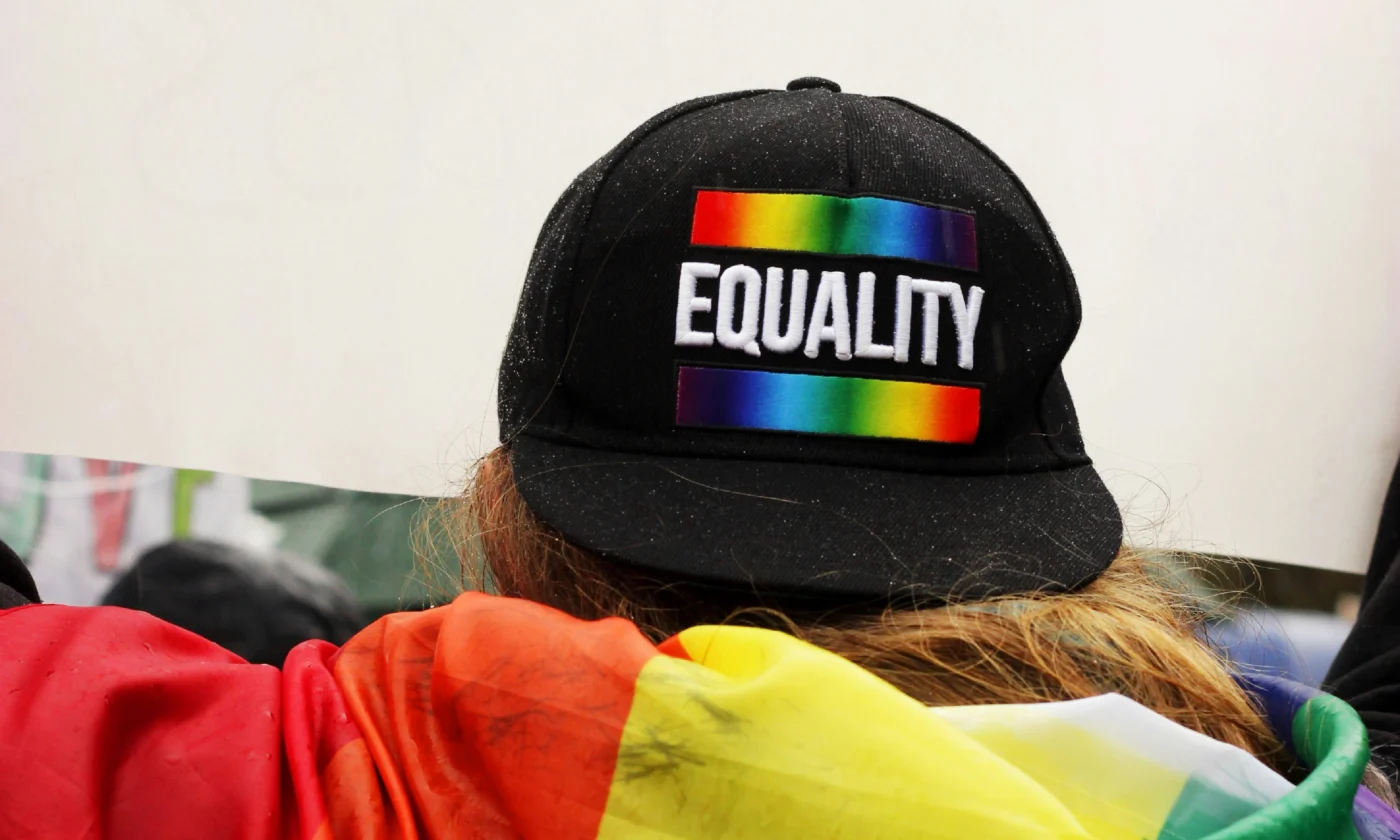 The back of a person cloaked in a rainbow flag and wearing a black baseball cap backwards. The word "EQUALITY" is printed on the front of the cap.