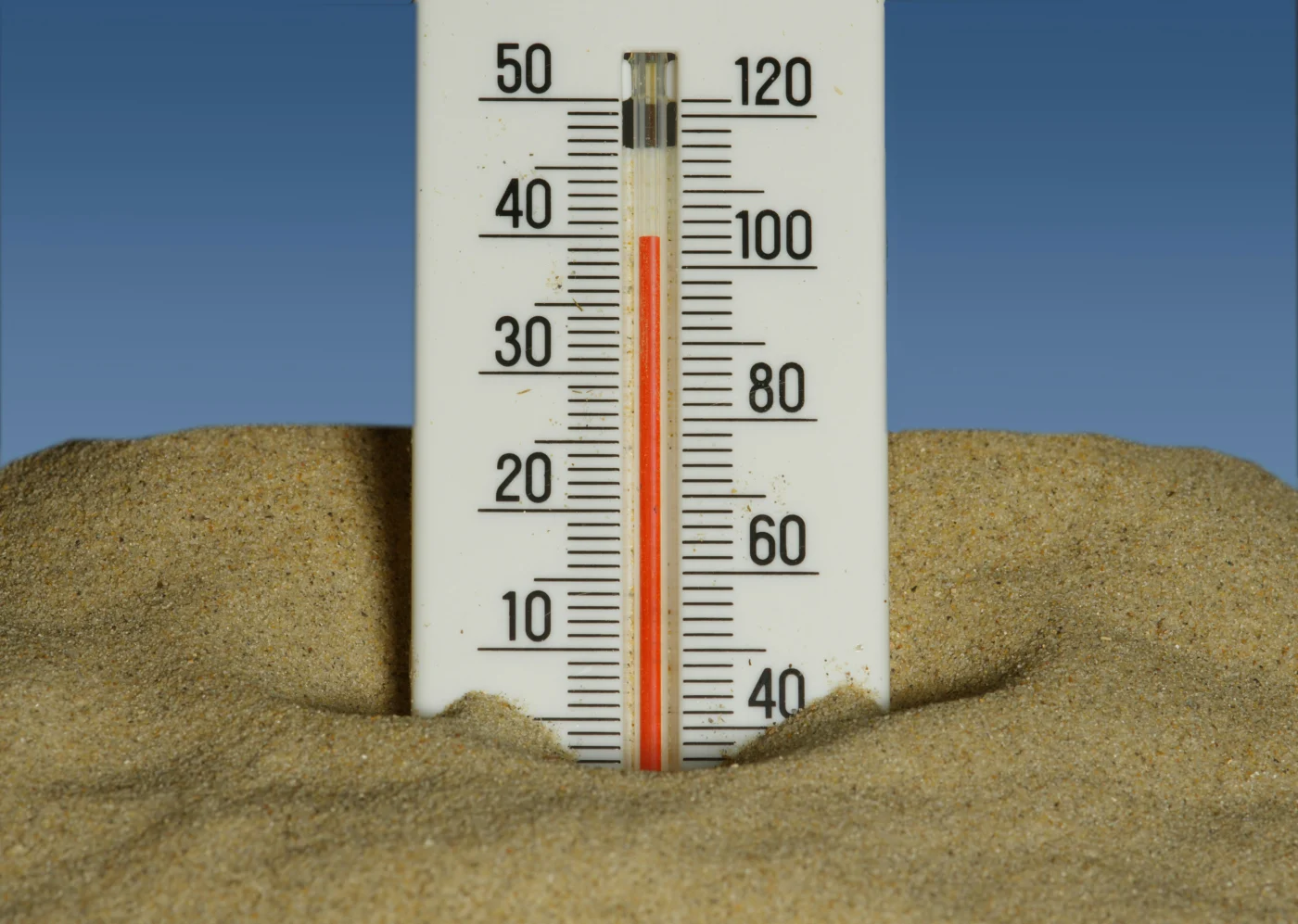 A thermometer half buried in sand in front of a blue sky.